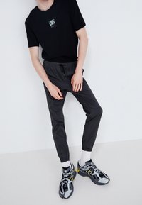 Černé bavlněné tričko s malým grafickým motivem, elastické tepláky v tmavě šedé barvě a vícebarevné tenisky New Balance s tlustými podrážkami a tkaničkami.