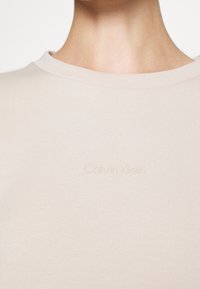 Primo piano di collo e parte superiore del torace che indossa una maglietta a girocollo beige con il logo "Calvin Klein" in rilievo al centro del tessuto.