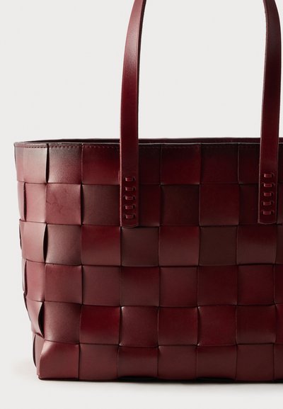 Μεγάλη δερμάτινη τσάντα ώμου μπορντό με φαρδιές πλεκτές λωρίδες και δύο ανθεκτικούς ιμάντες ώμου σε απλό φόντο.