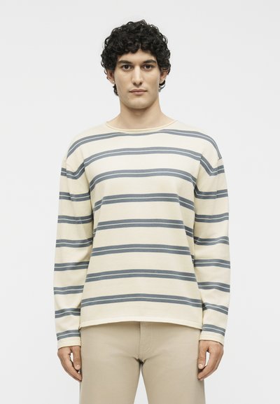 Jeune homme aux cheveux bouclés portant une chemise à manches longues crème et grise à rayures horizontales et un pantalon beige, debout devant un fond uni.