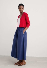 Rdeča, krojena jopica preko belega topa; mornarsko modra, visoko pasovana, nabranilka maksi krila, v kombinaciji z rjavimi usnjenimi sandali s trakovi.