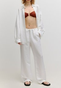 Weißes Leinen-Hemd in Übergröße, brauner Bikini mit goldenen Details, lockere weiße Hose und schwarze Flip-Flops. Glatte Textur, entspannte Passform.