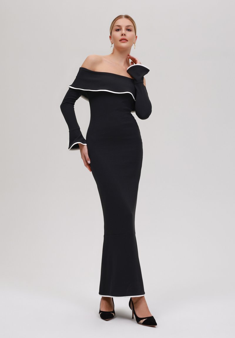 Svart off-shoulder klänning gjord av elastisk tyg med vit trim. Åtsittande bodice som fläker ut i botten. Modell som bär svarta klackar och har långa ärmar.