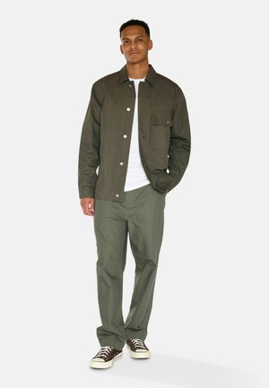 Olive green jacket with snap buttons and chest pocket, paired with matching olive pants and brown sneakers with white soles.