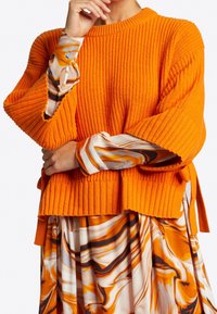 Pull orange chaud en maille côtelée avec un col rond et des manches larges, associé à une robe fluide à motifs dans des nuances d'orange et de crème.