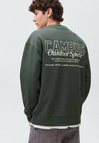 Felpa verde con scollo rotondo e polsini a costine, caratterizzata da testo bianco sul retro, inclusi "CAMPUS Outdoor Sports" e nomi di sport.