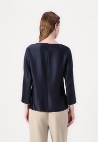 Kvinne med vått hår iført en langermet topp i marineblått og beige bukser, stående med ansiktet mot en hvit vegg, med ryggen helt synlig.