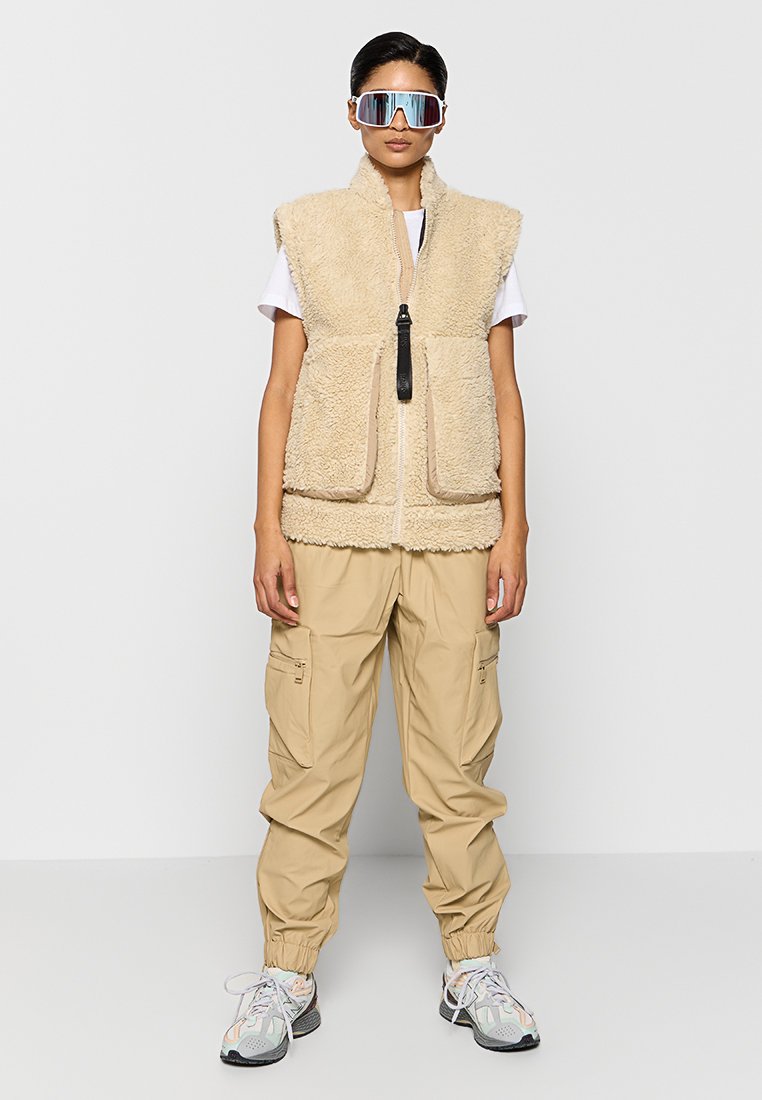 Donna che indossa un gilet in shearling beige, pantaloni cargo, sneakers bianche e grandi occhiali da sole riflettenti, in piedi su uno sfondo bianco.