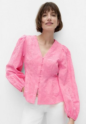 Kvinde iført en lyserød bluse med lange ærmer med blomstret broderi og knapper, kombineret med hvide bukser, stående mod en ensfarvet hvid baggrund.