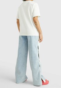 Bílá bavlněná tričko s krátkým rukávem, doplněné světle modrými džíny s širokými nohavicemi a bočními rozparky, nošeno s růžovými gumovými sandály.