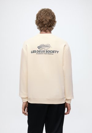 Personne aux cheveux bouclés portant un sweat crème avec l'inscription « LES DEUX SOCIETY PASSION FOR PEOPLE » et un graphique représentant un globe dans le dos.