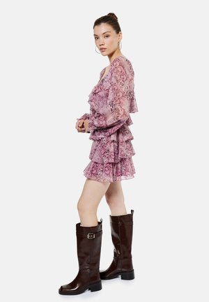 Nuori nainen vaaleanpunaisessa kerroksellisessa röyhelömekossa ja korkeissa tummanruskeissa nahkasaappaissa, esittäytyen profiilissa valkoista taustaa vasten.