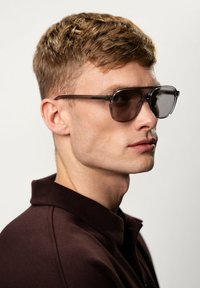 Zonnebril met een transparant grijs montuur en donkere glazen, met dunne zwarte steunen en een rechthoekige vorm, gedragen door een model.
