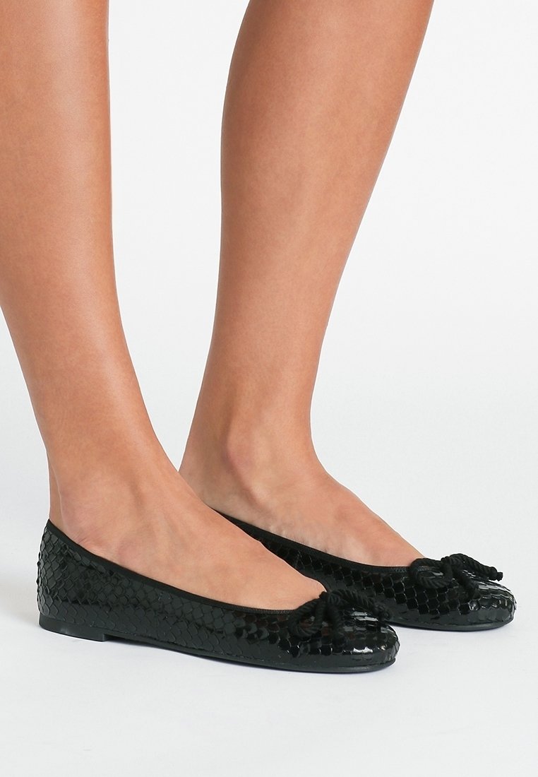 Pieds de femme portant des ballerines noires texturées avec de petits nœuds sur fond blanc.