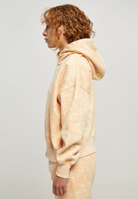 Hoodie v broskovej farbe s uvoľneným strihom, ktorý má textúrovaný, mramorovaný vzor a priestrannú kapucňu. Ramená a lem sú rebrované.