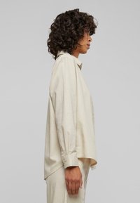 Beige skjorte med knapper og lange ermer, krage og bølget nederkant; laget av et mykt, lett stoff; minimalistisk design uten mønstre.