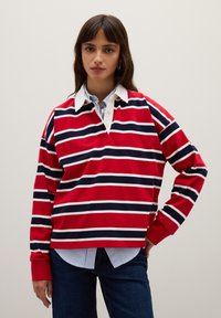 Rode langegsmouf polo met witte en marineblauwe horizontale strepen, met een witte kraag en een gedeeltelijke knoopsluiting, gedragen over een blauw gestreept shirt.