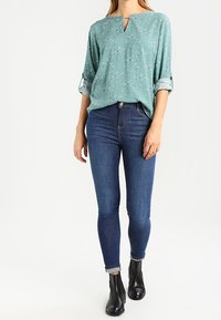 Blusa teal con stampa floreale e scollatura a chiave, abbinata a jeans skinny blu scuro e stivaletti neri.