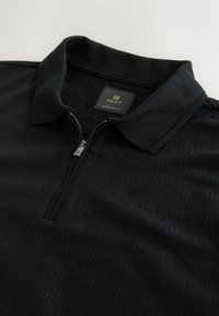 Svart pikétröja med krage som kan dras upp, med ett strukturerat geometriskt mönster och en tagg märkt "NEXT REGULAR FIT."