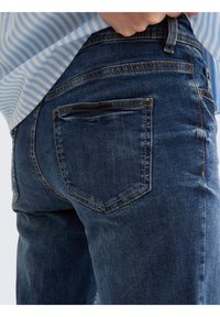 Los pantalones vaqueros de denim azul oscuro presentan un diseño de cinco bolsillos con costuras visibles y un corte recto. La tela tiene una textura suave y ligeramente desgastada.