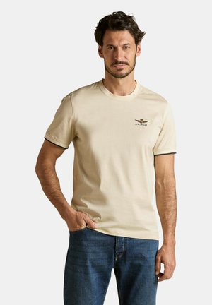 Uomo con capelli corti e scuri e barba curata, che indossa una maglietta beige a maniche corte con un piccolo logo e jeans blu, in piedi con una mano in tasca.