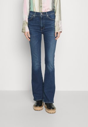 Personne portant un jean bootcut taille haute bleu foncé, un t-shirt rayé multicolore à manches longues, et des bottes noires épaisses avec semelles couleur tan.