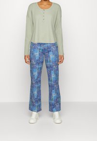 Ljusgrön långärmad topp med knappar, i kombination med blå mönstrade byxor som har olika texturer och färger, avslutat med vita sneakers.