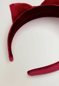 Punainen samettinen hiuspanta, jossa on kissankorvat ja pieni satiinipanta valkoista taustaa vasten.