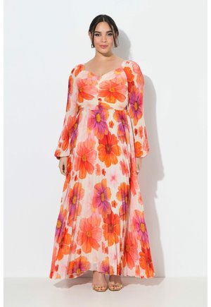 Eine Frau steht da und trägt ein langes, plissiertes Kleid mit großen orangefarbenen und purpurfarbenen Blumenmustern sowie durchsichtige lange Ärmel, kombiniert mit goldenen Sandalen mit offenem Zehenbereich.