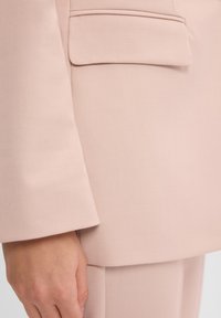 Zbliżenie na materiał różowej marynarki, z gładką teksturą, pojedynczą kieszenią i wyraźnie uformowanymi rękawami. Dopasowany krój z minimalistycznymi elementami designu.