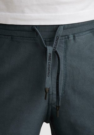 Primo piano di pantaloni della tuta grigio scuro strutturati con coulisse nere con il marchio "PME Legend" e una camicia bianca infilata leggermente sopra la vita.