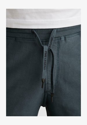 Nahaufnahme dunkelgrauer, strukturierter Sweatpants mit schwarzen „PME Legend“-Marken-Kordeln und einem weißen Hemd, das leicht über dem Bund steckt.