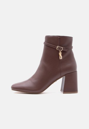 Brown leather ankle boot with block heel, pointed toe, and decorative gold buckle and charm strap around the ankle.