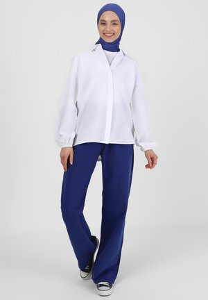 Camicia bianca con bottoni e maniche a palloncino abbinata a pantaloni larghi blu navy. Il modello indossa un hijab blu e sneakers nere.