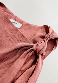 Vaaleanpunainen mekko, jossa on lehtikuvioinen tekstuuri, jossa on solmittu solmu yksityiskohta hihassa ja etiketti, jossa lukee "NEXT". Silkkinen kankaan pinta.