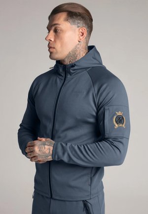 Man met tatoeages op nek en hand, gekleed in een donkerblauwe hoodie met rits en een embleem op de linker mouw, staand tegen een effen achtergrond.