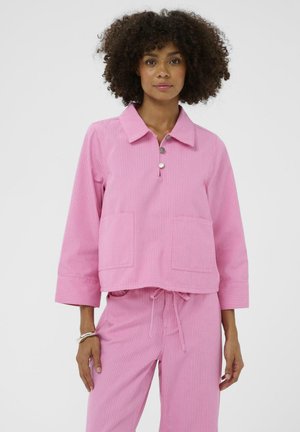 Ružičasti pulover od kostimnog pana s ovratnikom, rukavima tri četvrtine, dva prednja džepa i detaljem s gumbima. Stiliziran s odgovarajućim ružičastim hlačama.