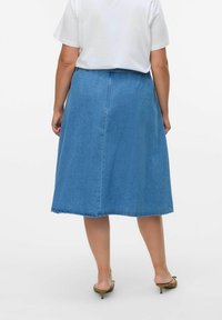 Gonna midi in denim con una forma leggermente a trapezio, di colore blu, orlo grezzo e cucitura centrale sul retro, abbinata a una t-shirt bianca e sandali.