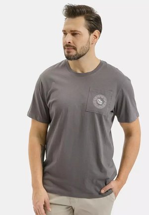 Mężczyzna z ciemnymi włosami i brodą, ubrany w szarą koszulkę z okrągłym logo na kieszeni na piersi, beżowe spodnie, stojący z jedną ręką w kieszeni.