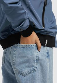 Veste bomber bleu clair avec poignets élastiques noirs, fermeture éclair à l'avant et poche latérale. Portée avec un jean en denim bleu clair avec détails de poche visibles.