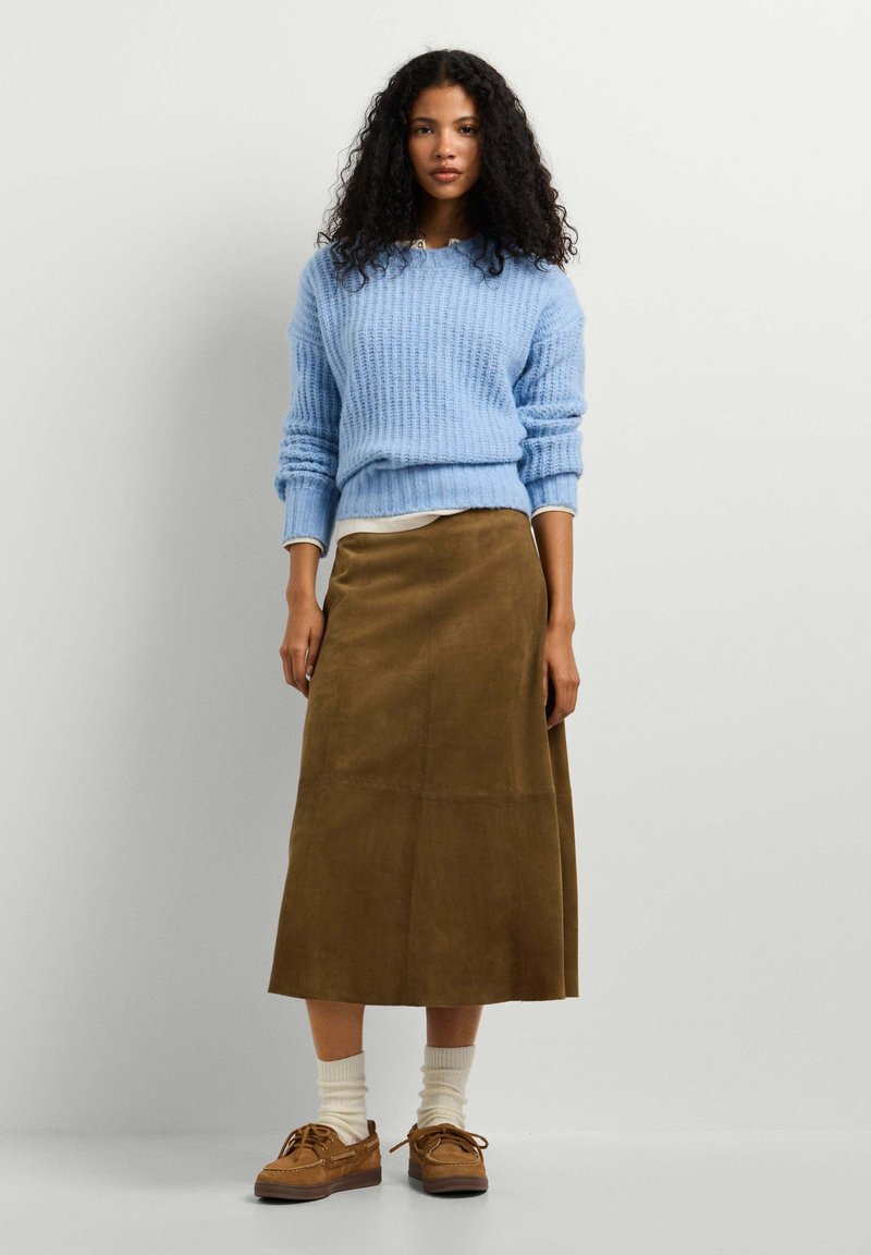 Femme aux cheveux bouclés portant un pull en maille bleu clair, une jupe midi marron, des chaussettes crème et des chaussures marron à lacets, debout devant un fond uni.