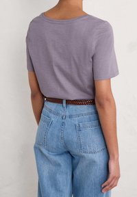 Persona che indossa una camicia lavanda a maniche corte infilata in jeans a vita alta di colore blu chiaro, con una cintura intrecciata marrone, vista da dietro.