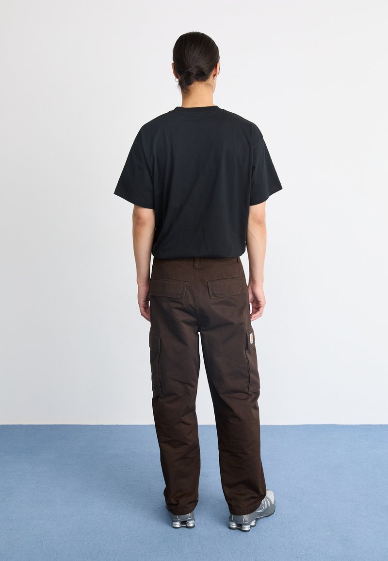 Zwarte T-shirt en bruine cargo-broek met zijzakken, gemaakt van stevig materiaal. Model staat op een blauwe vloer tegen een witte achtergrond.