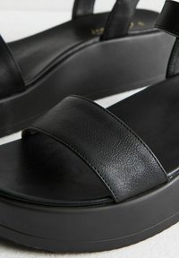 Svarta plattformsandaletter med breda läderremmar och tjocka vadderade sulor på en ljus bakgrund.