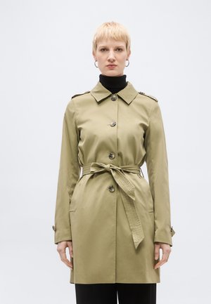 Person iført en beige trenchcoat med bælte over en sort rullekrave, stående mod en ensfarvet lys baggrund og vendt fremad.