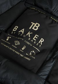 Indvendig label i sort pufferjakke med "BAKER" logo i guld, samt ikoner der angiver vandmodstand, holdbarhed, varme og genanvendt fyld.