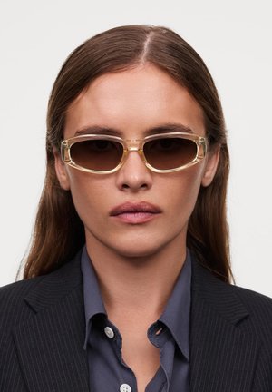 Solsikkers med en gjennomsiktig gul ramme, oval form og brune linser. Brukt med en mørkstripet blazer og blå skjorte.