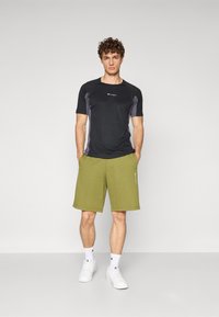 Svart träningsskjorta med grå accenter, ihop med olivgröna shorts. Vita sneakers och ankelsockor fulländar outfiten.