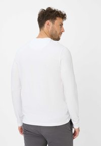 Hvid langærmet skjorte med rund hals, glat tekstur og tætsiddende design, parret med grå bukser, vist fra ryggen.