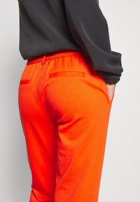 Rood-oranje joggingsbroek met een elastische tailleband, voorzien van twee achterzakken en een gladde textuur. Gecombineerd met een grijs shirt, met details op de mouwen.